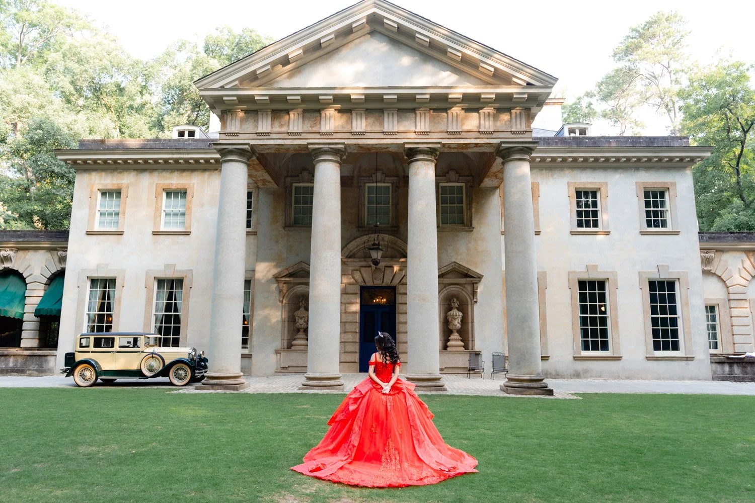 A woman in a red ball gown standing on a lawn in front of a large, historic mansion with tall columns and a vintage car parked nearby.