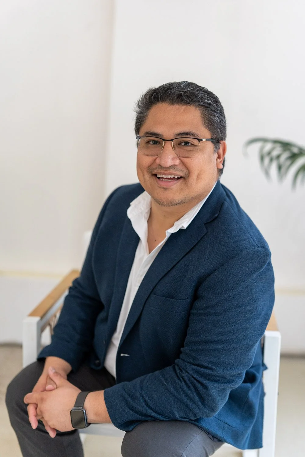 A man with glasses and dark hair, wearing a navy blue blazer and white shirt, sitting on a white chair in a bright room with a neutral background and a potted plant.
