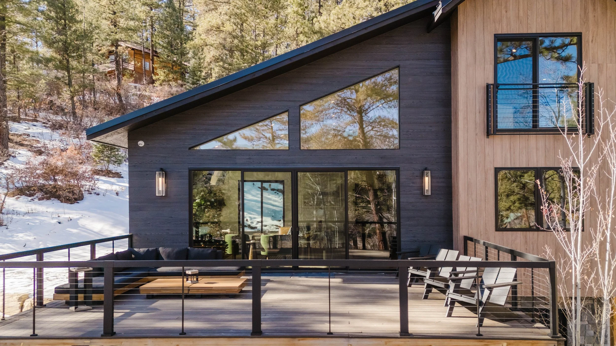 Modern two-story house with large windows and a spacious outdoor deck, situated in a snowy, wooded area.