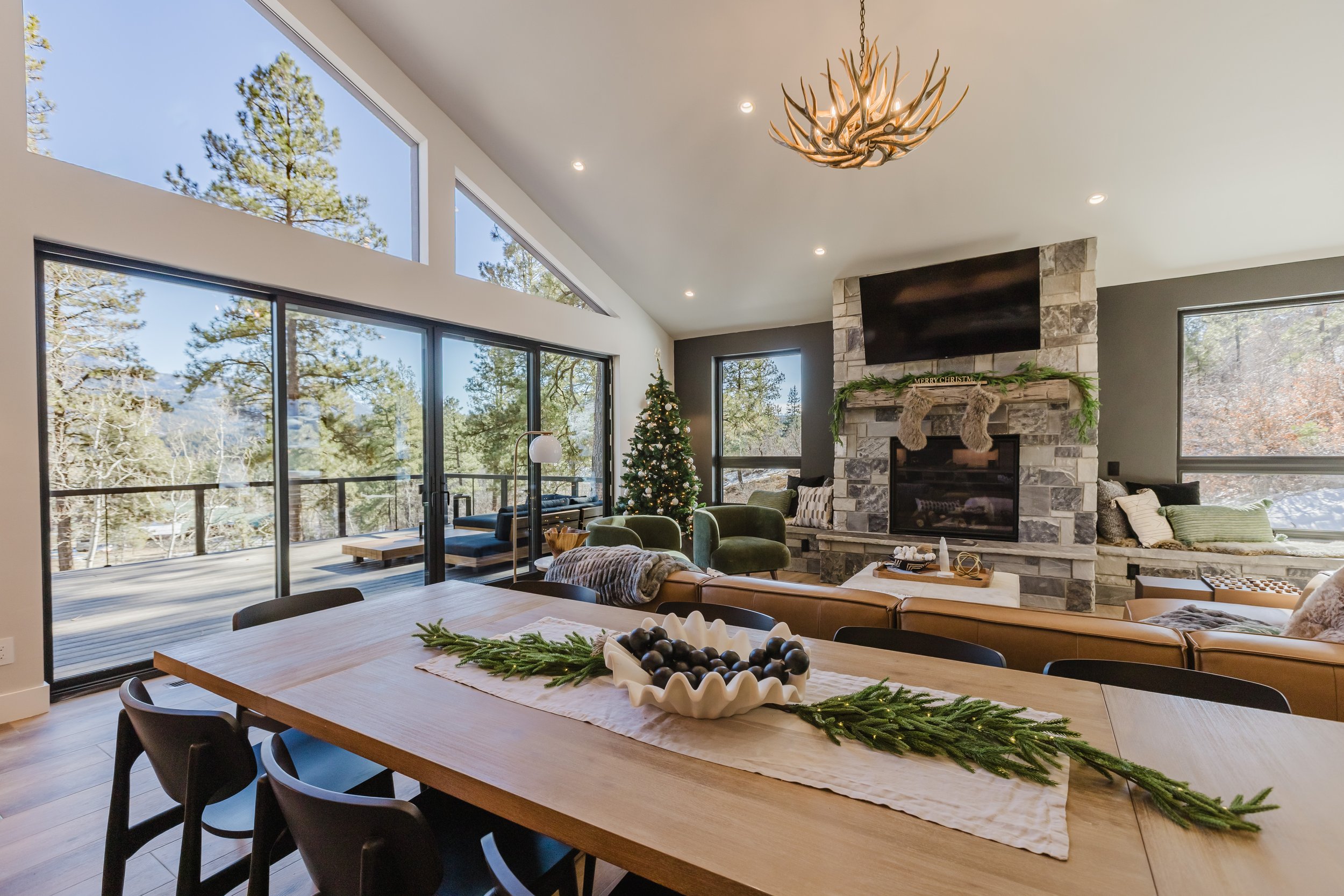 Interior of a modern living room decorated for Christmas with a Christmas tree, stockings, and a festive mantle, featuring large windows showcasing a forested landscape outside.