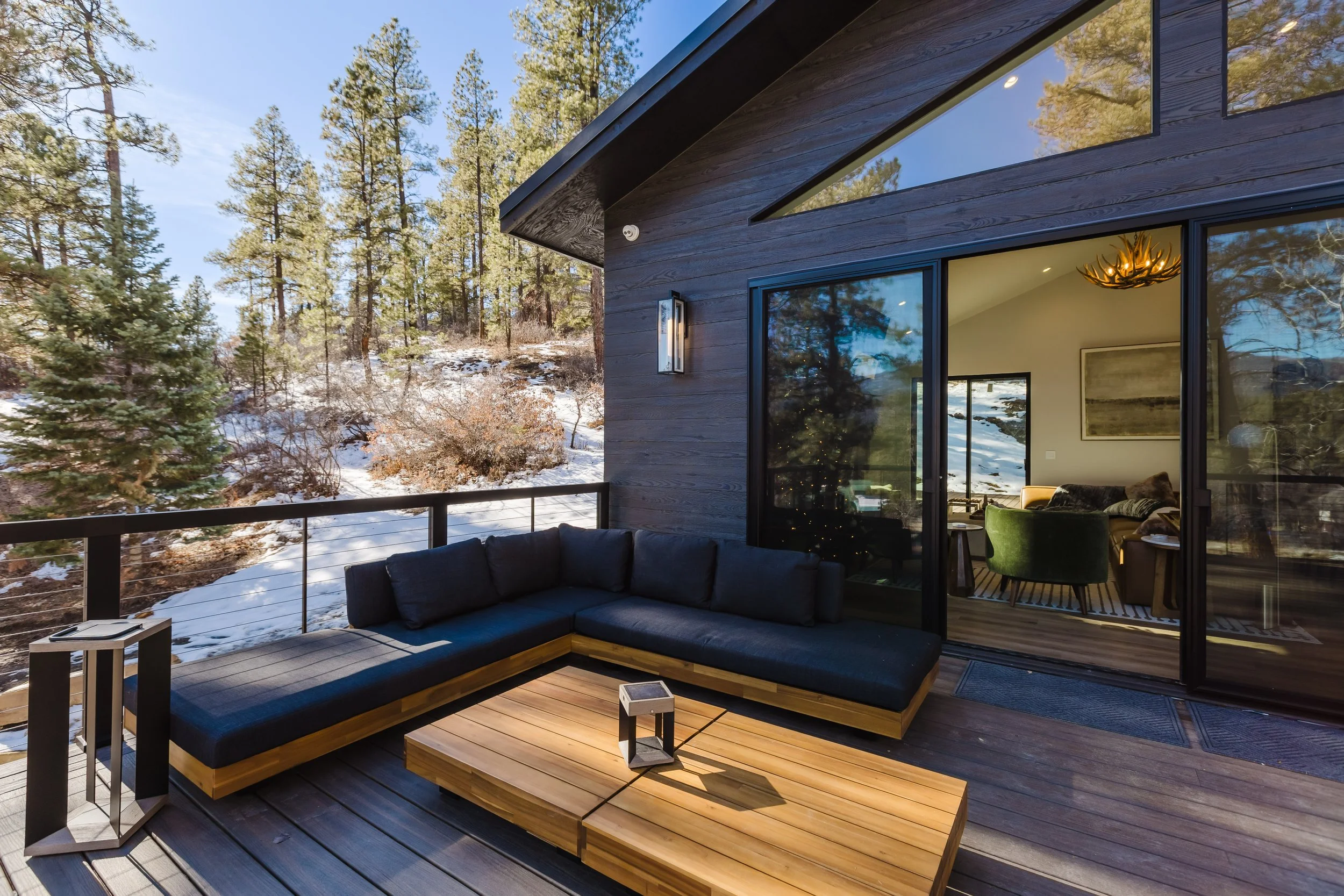 Modern house with large glass windows and a wooden deck balcony overlooking a snowy forest landscape, with trees and blue sky visible.