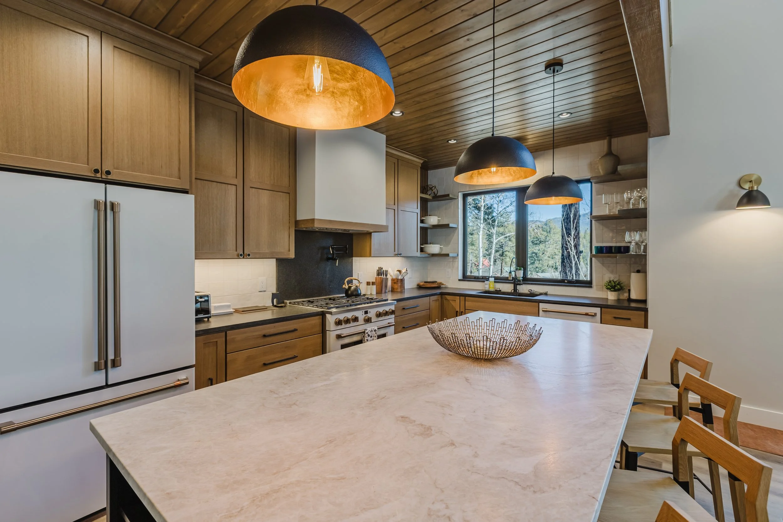 A modern kitchen with wooden cabinets, a white fridge, a marble countertop island, black and copper pendant lights, a window with a view of trees, and various kitchenware.