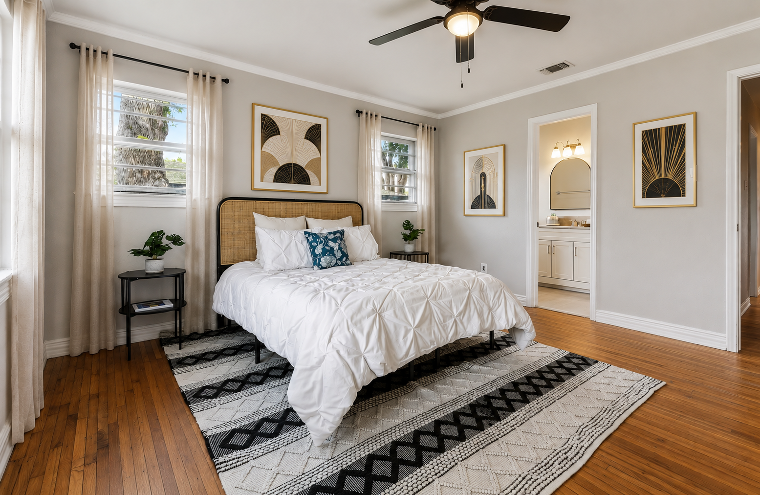 A cozy bedroom with a large bed, white bedding, and four pillows, two large white pillows and one decorative blue pillow with white floral pattern. There are two black nightstands with potted plants on each side of the bed. The room has light gray walls, hardwood floors, and white curtains on the windows. Decor includes framed abstract art on the walls. A ceiling fan is mounted on the ceiling, and a bathroom is visible through an open doorway.