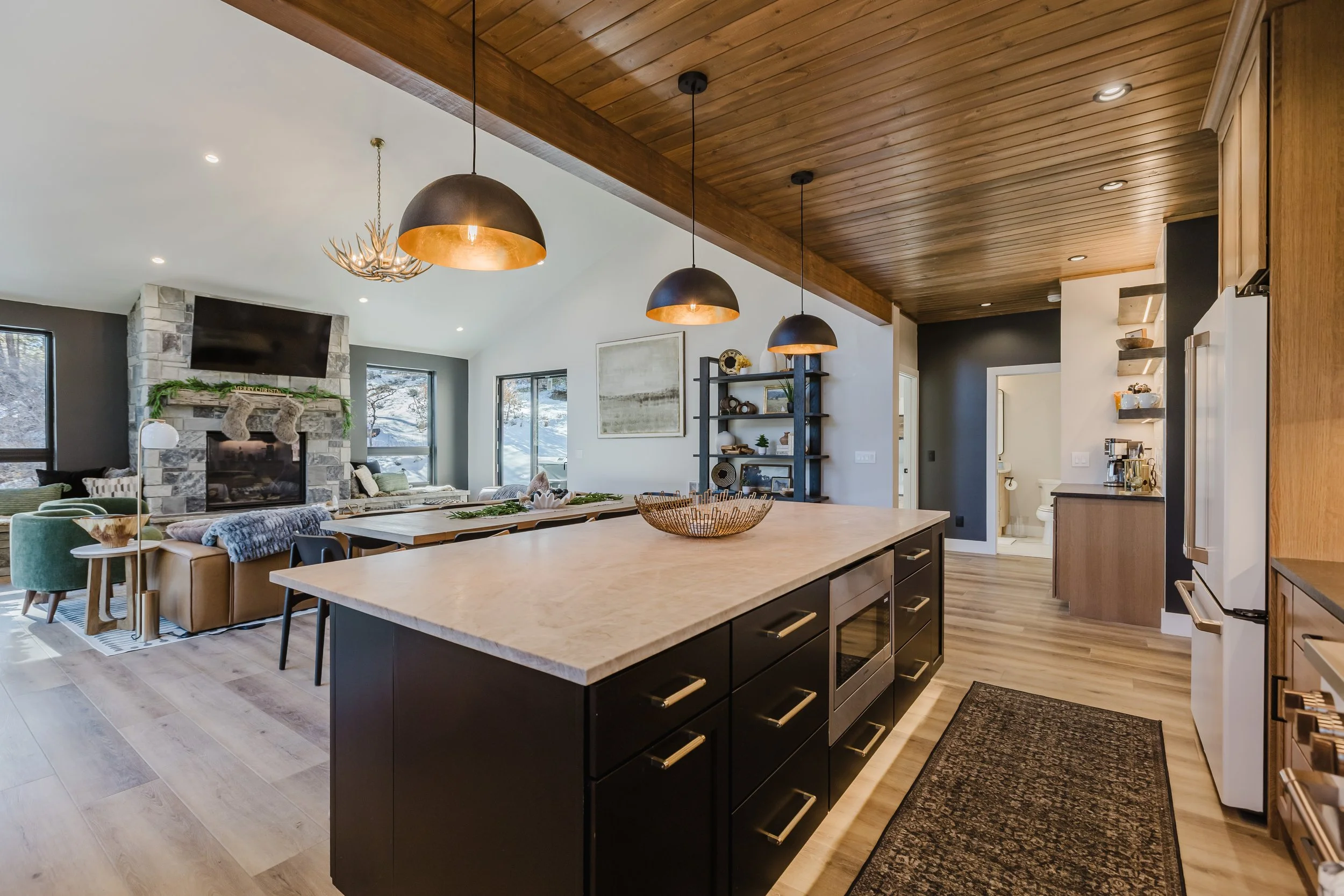 Modern open-concept kitchen and living room with wooden ceiling, black island, fireplace, and large windows showing snow outside.