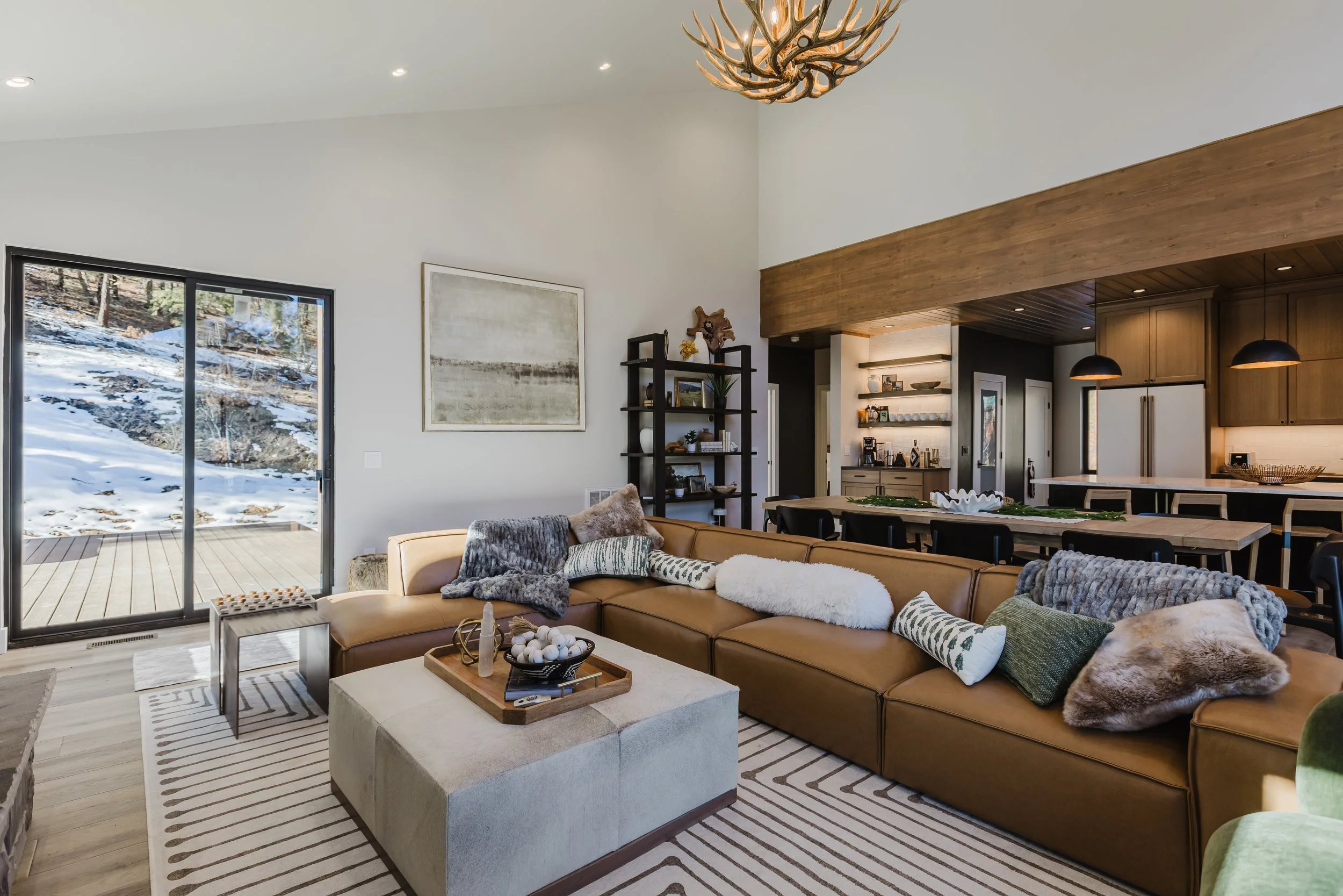 Living room with a tan sectional sofa, white ottoman, and decorative pillows in a modern house. Large sliding door shows a snowy outdoor landscape. Art and shelving decor on the wall, wooden beamed ceiling, and a kitchen area with hanging black lamps.