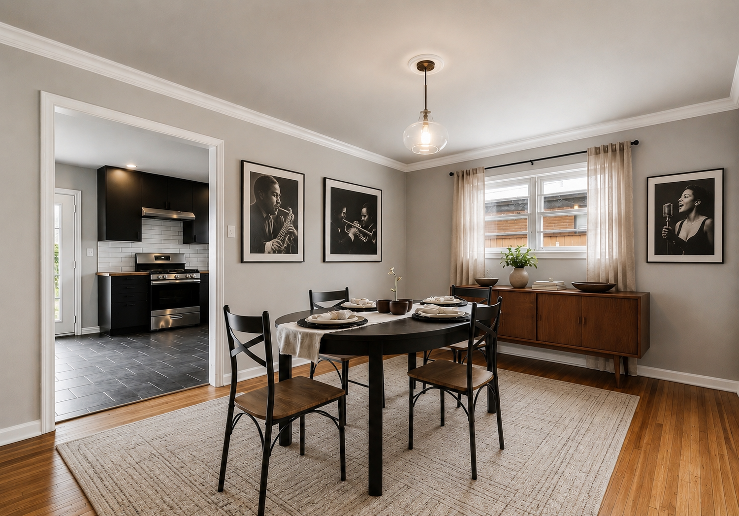 Dining room with round black table, four chairs, window with curtains, wall art of jazz musicians, sideboard, houseplant, and a view into the kitchen with black cabinets and stainless steel stove.