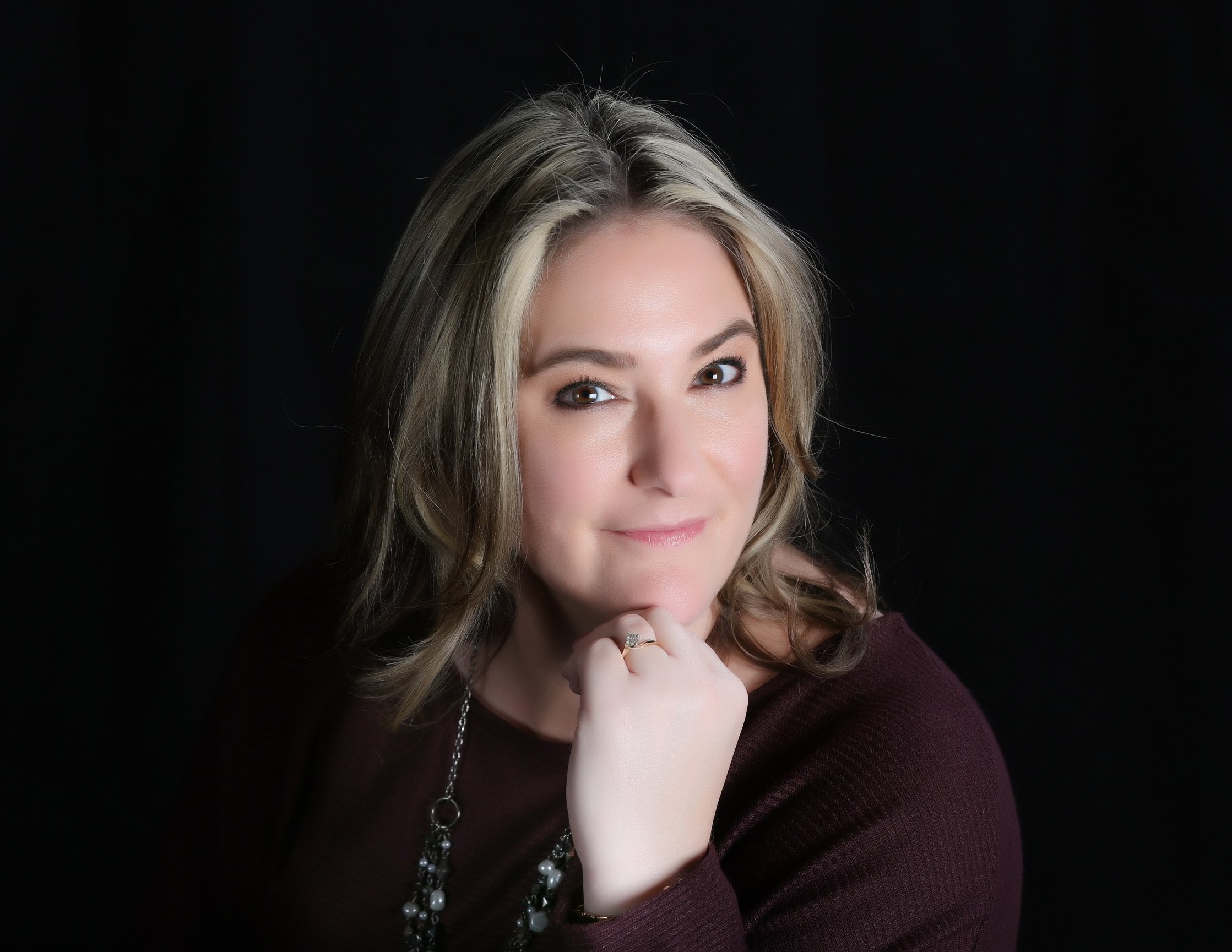 Photo of a woman with blonde hair styled in loose waves, wearing a dark maroon top, a pearl necklace, and a ring with a prominent stone, against a black background.