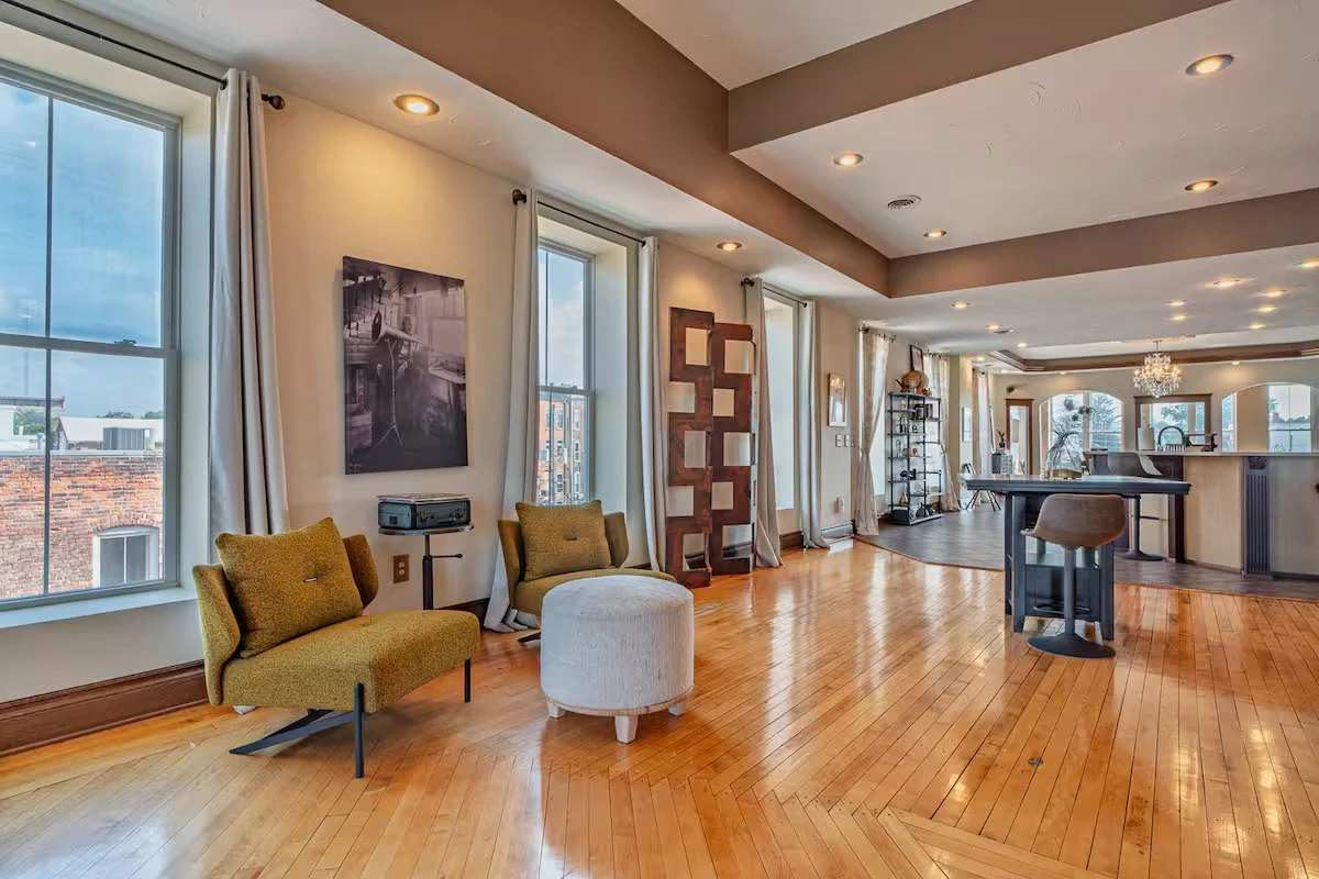 Living room with large windows, hardwood floors, modern furniture including chairs, ottoman, bar stool at a breakfast bar, and a decorated open space with a chandelier and shelves. The Stewart Penthouse Living Room.