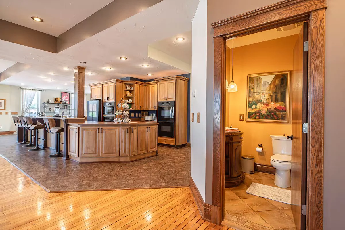 Kitchen with wooden cabinets, island, and bar stools next to a bathroom with a toilet and framed painting.