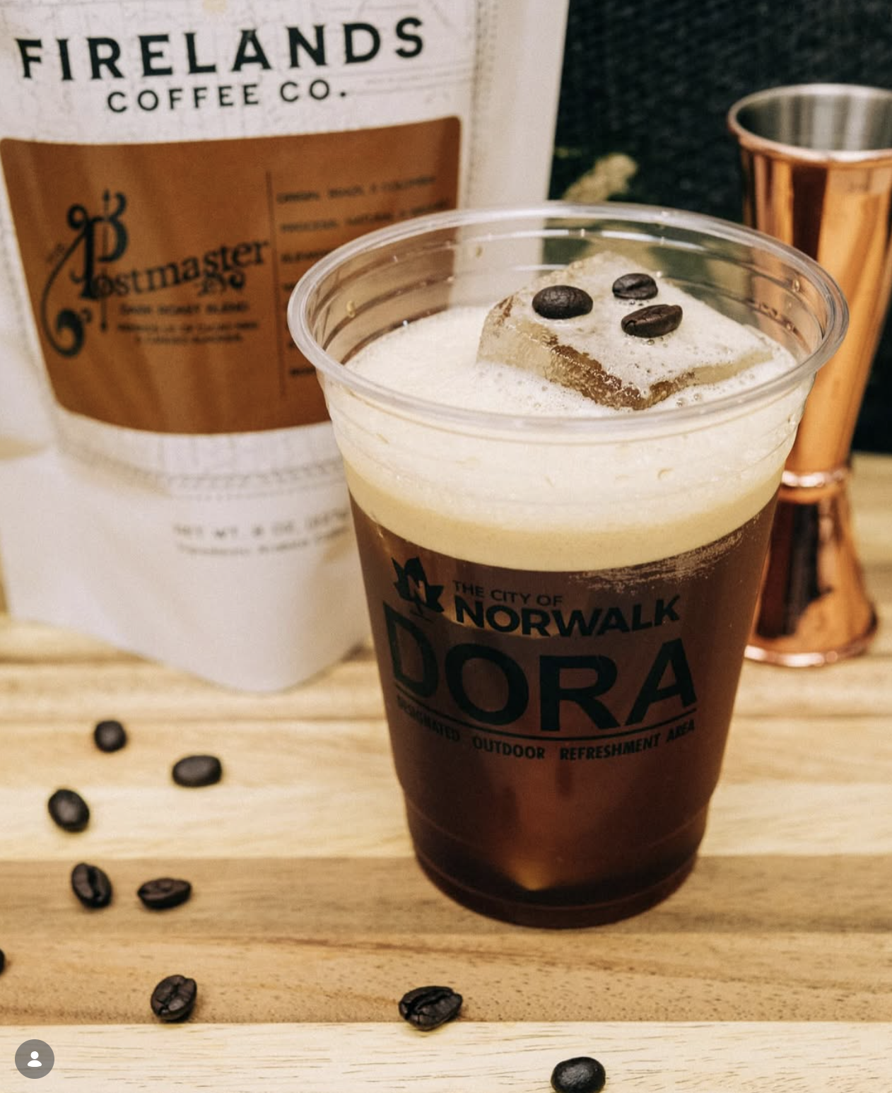 A plastic cup of dark iced coffee with foam and coffee beans on top, sitting on a wooden surface with scattered coffee beans around it. In the background, there's a package of Firelands Coffee Co. Postmaster coffee, a copper-colored cup, and a dark textured surface.