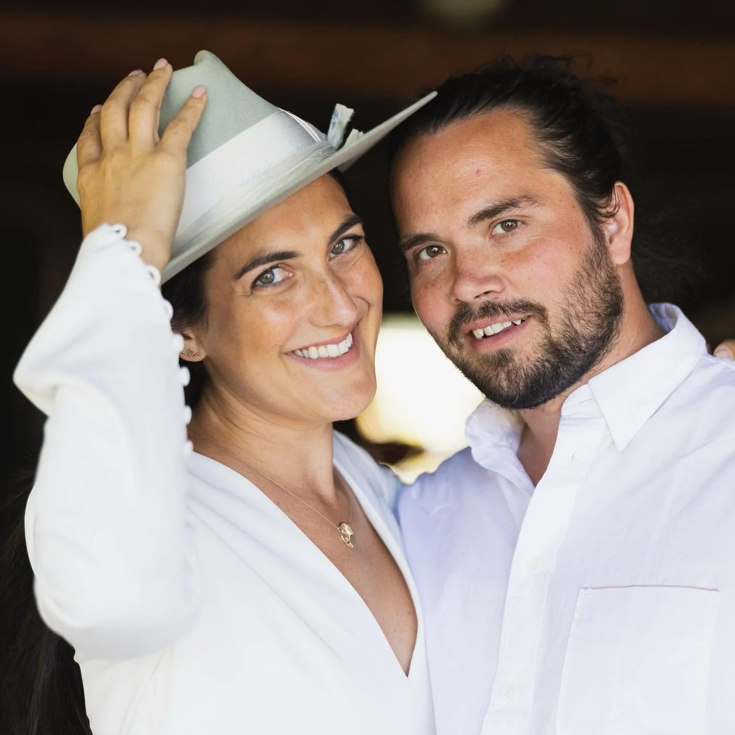 A smiling woman in a white shirt adjusting a hat on her head, standing next to a smiling man with a beard in a white shirt, both looking at the camera.