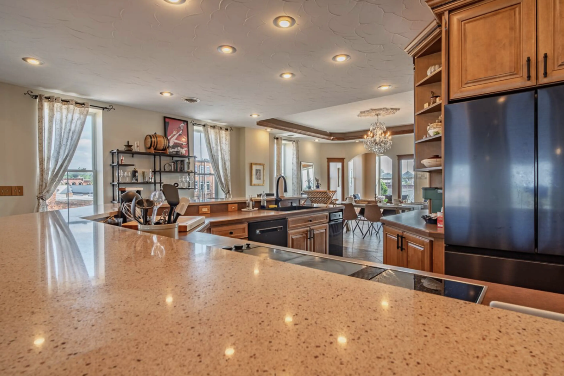 View of a spacious kitchen and dining area with wooden cabinets, a chandelier, large windows with curtains, and a granite countertop.
