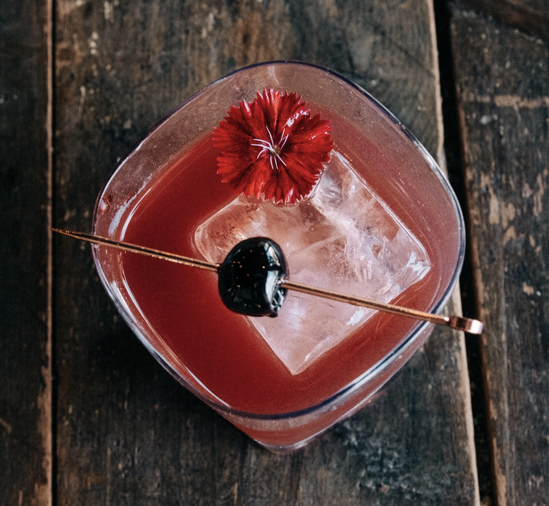 A pink cocktail in a glass with ice, garnished with a red flower and a skewered black cherry, placed on a rustic wooden surface.