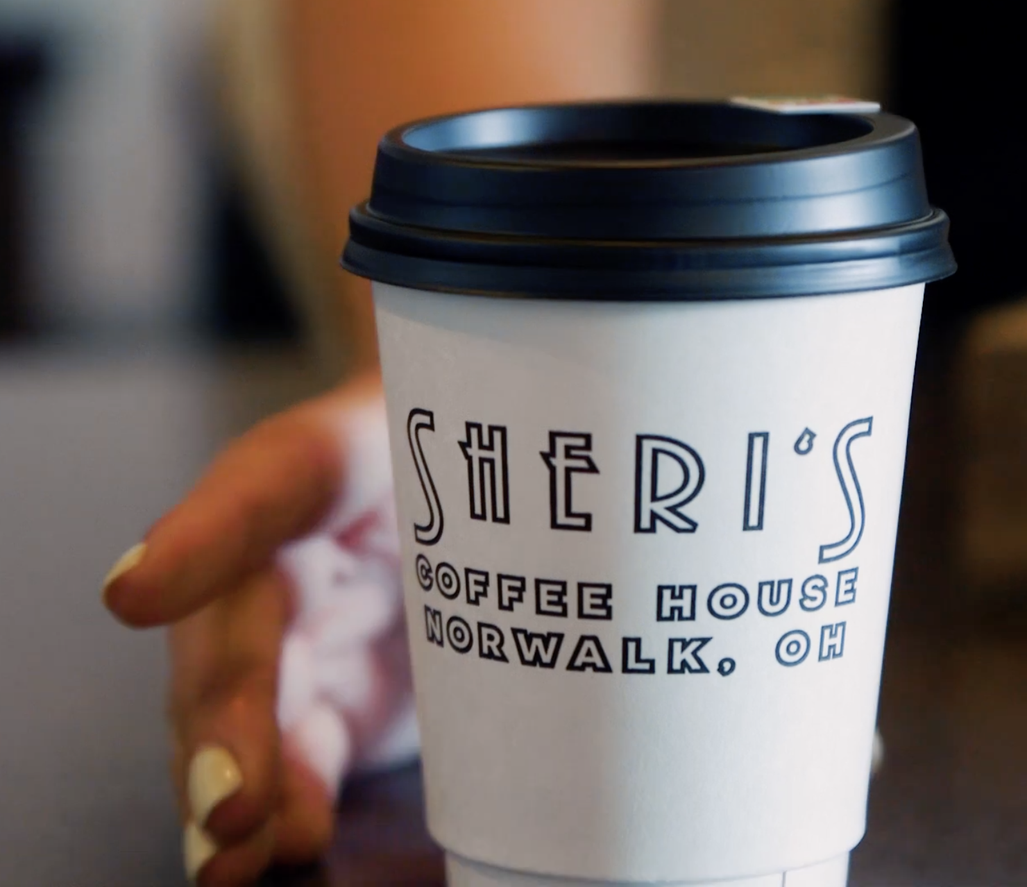 A hand holding a white paper coffee cup with a black plastic lid, printed with text that says 'SHERI'S COFFEE HOUSE, NORWALK, OH'.