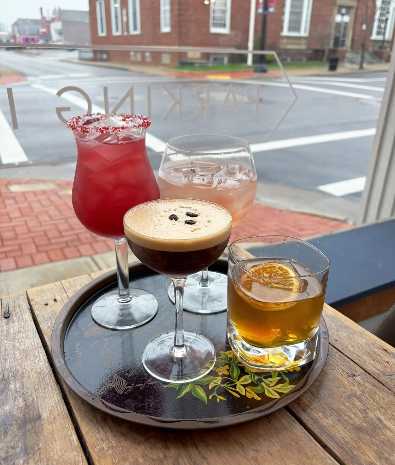 A tray with five cocktails and drinks placed on a wooden table inside a café, with a view of a street and brick buildings outside the window.