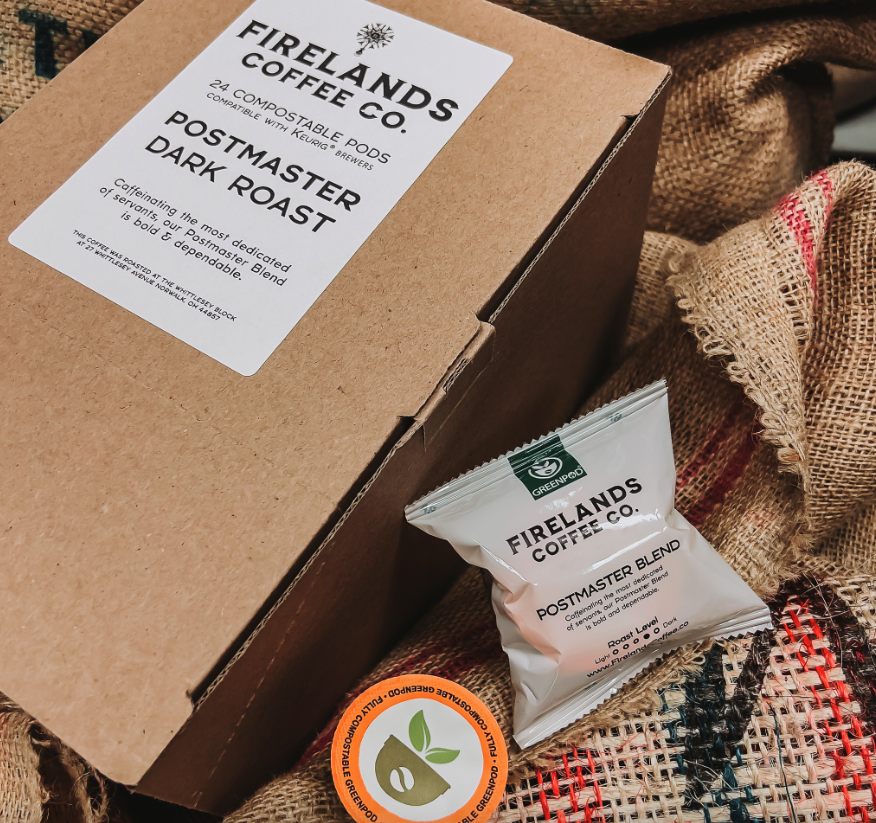 Box of Firelands Coffee Co. Postmaster Dark Roast coffee pods with a single pod and a compostable sticker, set on a woven fabric surface.