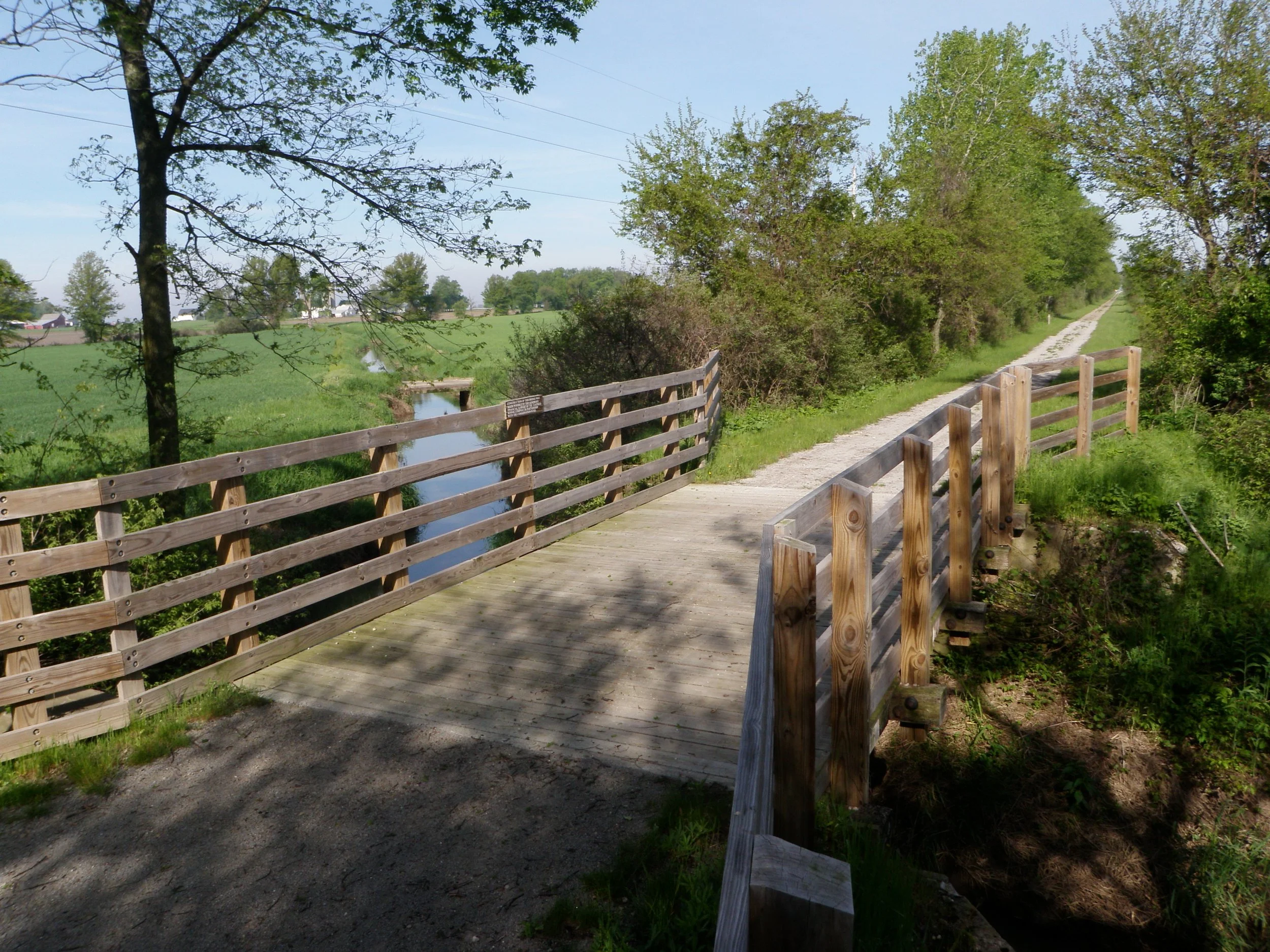 A rural scene with a dirt pathway and a wooden railing on the right side. The pathway runs alongside a small waterway, surrounded by lush green trees and open fields in the background under a clear blue sky.
