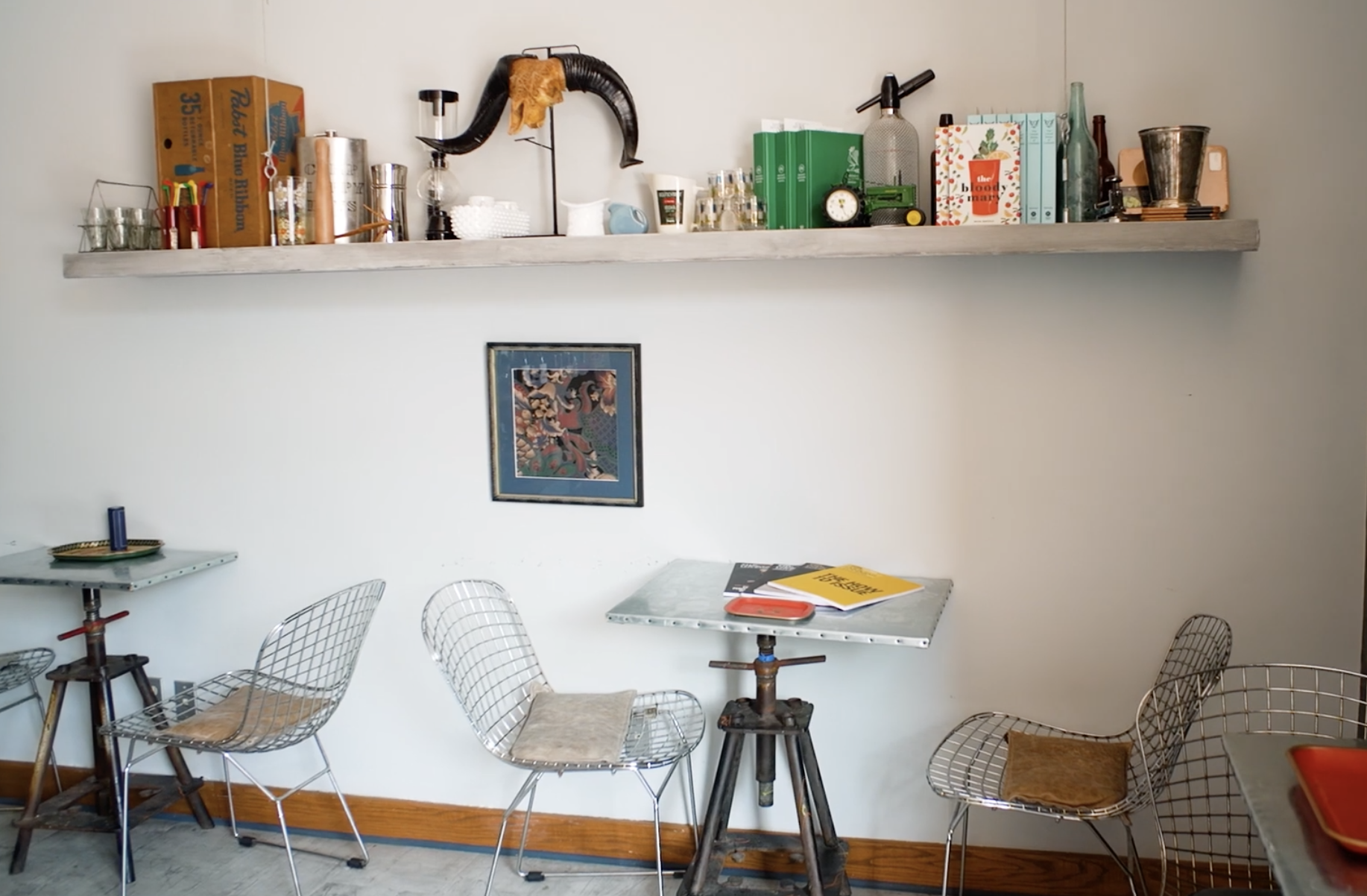 Interior of a modern café or dining area with metal wire chairs, tables, a wall shelf with various decorative objects, and a framed artwork on the wall.
