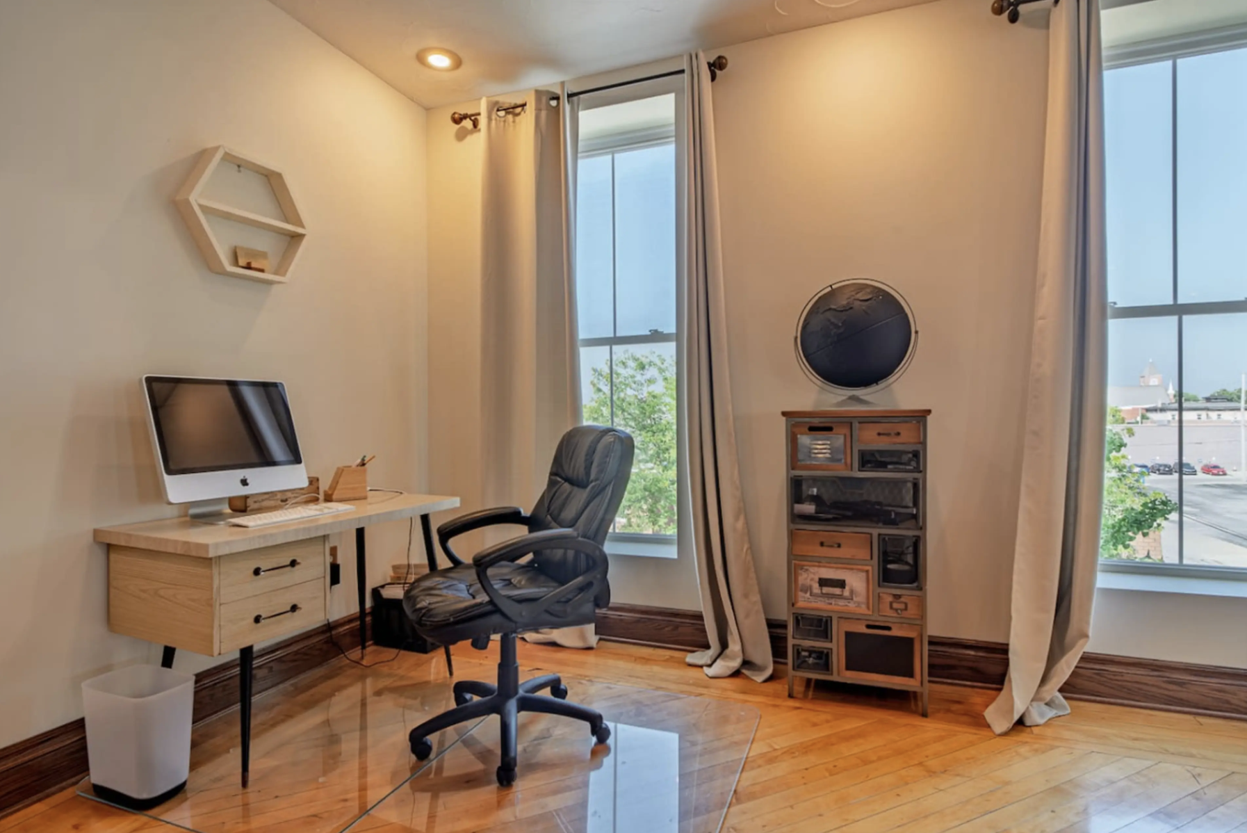 A home office with a wooden desk holding an iMac computer, a black office chair, and a transparent desk mat. The room has large windows with white curtains, and a small wooden cabinet with multiple drawers and transparent compartments stands against 