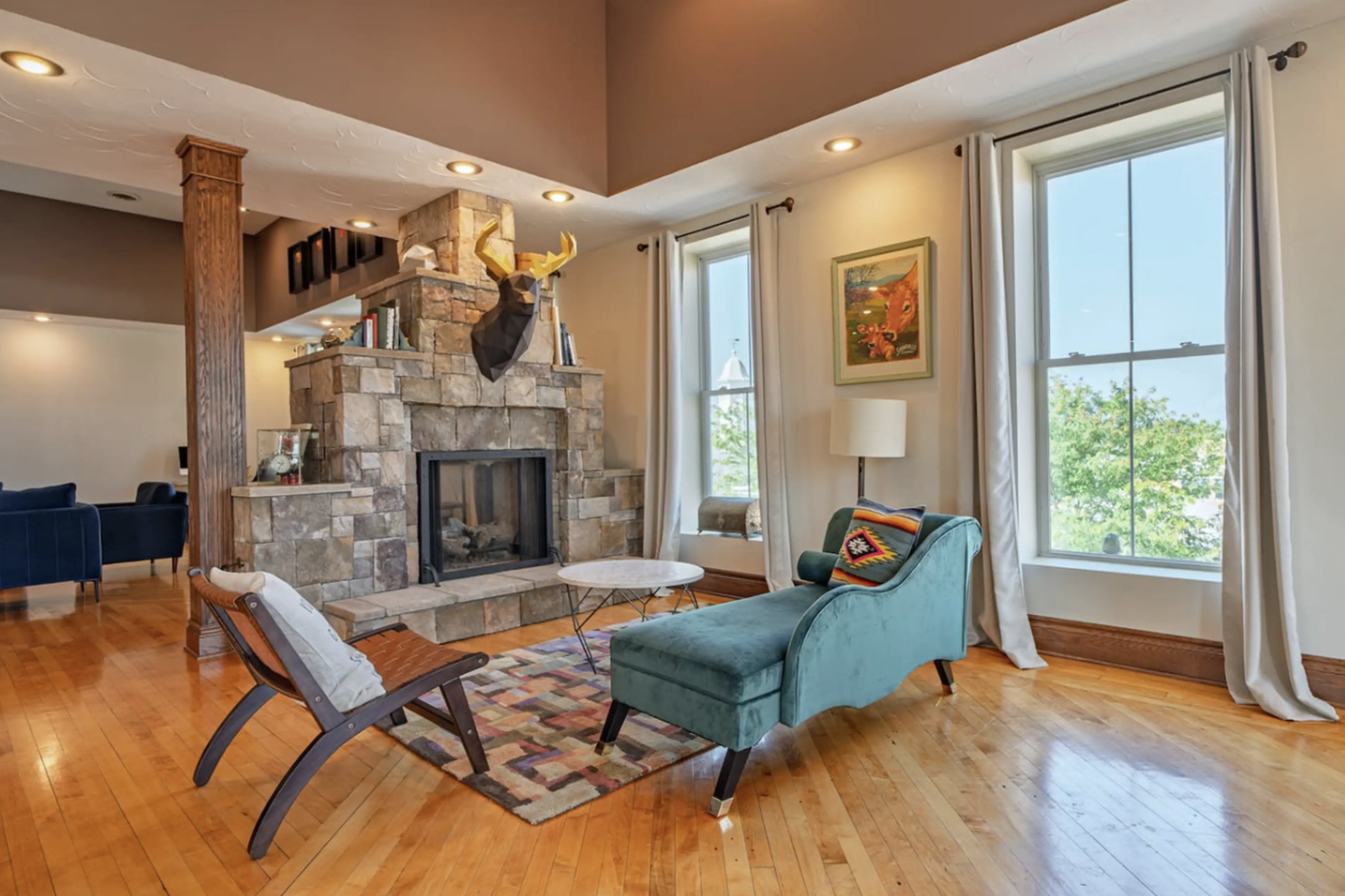 Living room with a stone fireplace, blue armchair, wooden rocking chair, large windows with white curtains, colorful rug, and various decorations including a mounted deer head, framed artwork, and a floor lamp.