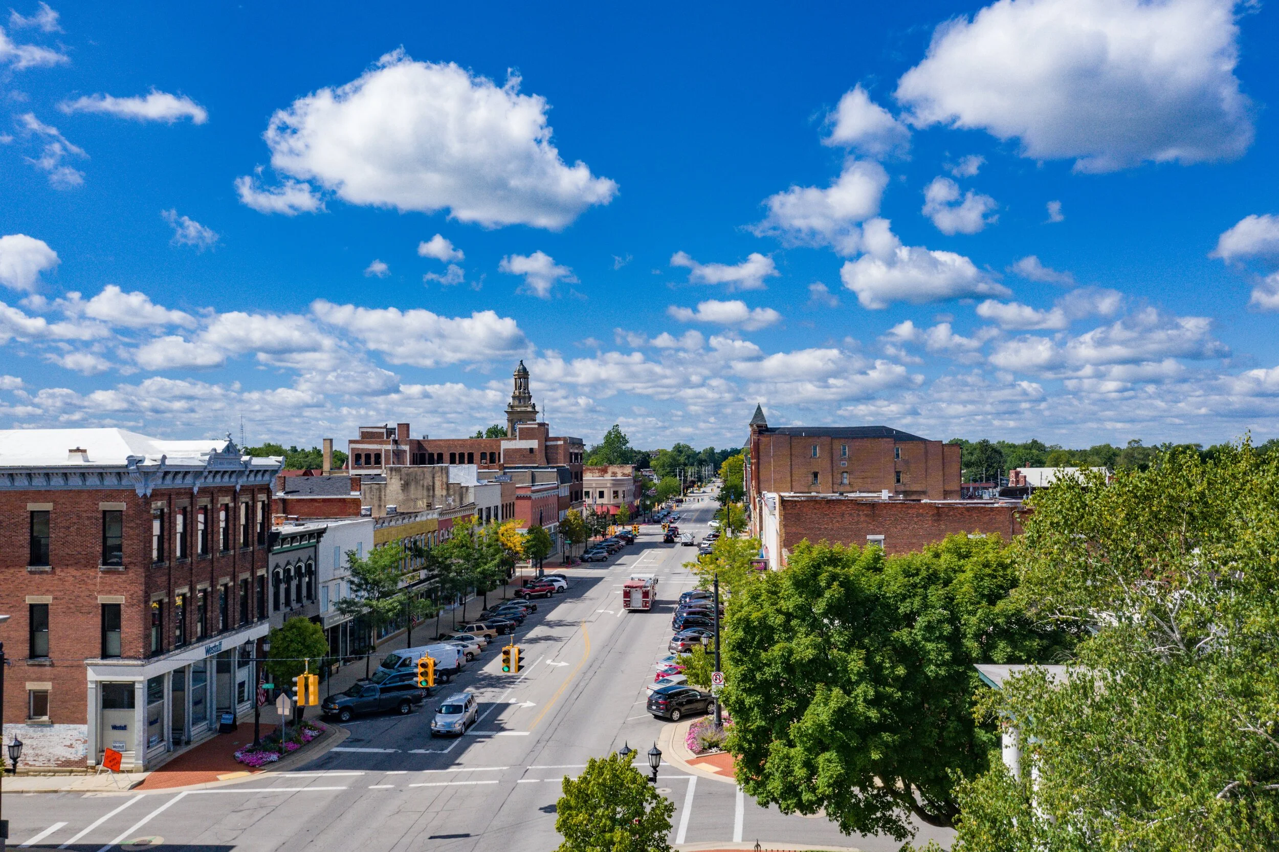 A small American town seen from an elevated view, with brick buildings lining a main street, cars parked along the street, trees, and a blue sky with scattered clouds.