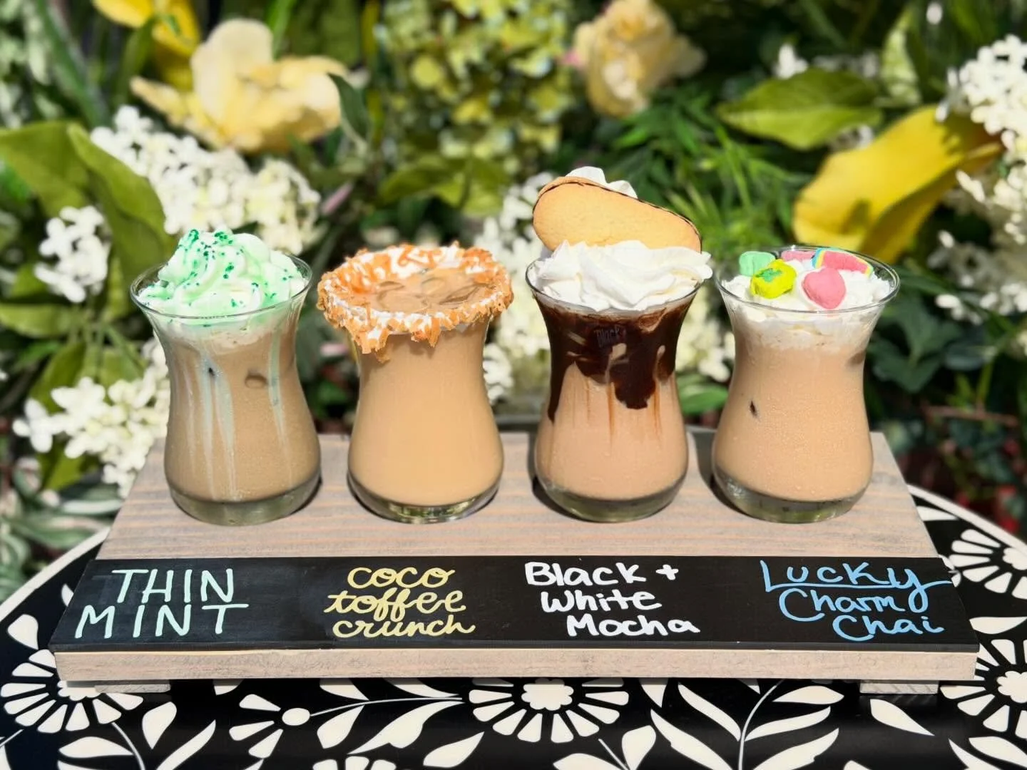 Four glasses of flavored coffee drinks with whipped cream and toppings arranged on a wooden board with names written in colorful chalk, placed outdoors with green foliage in the background.