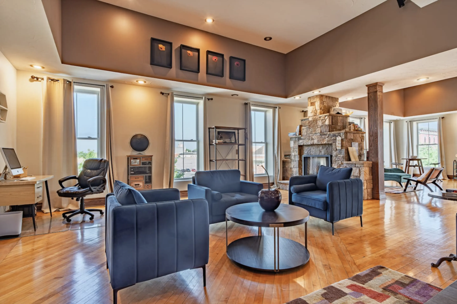 Spacious living room with hardwood floors, blue armchairs, a round black coffee table, a stone fireplace, or three large windows with beige curtains, and various furniture and decorations.