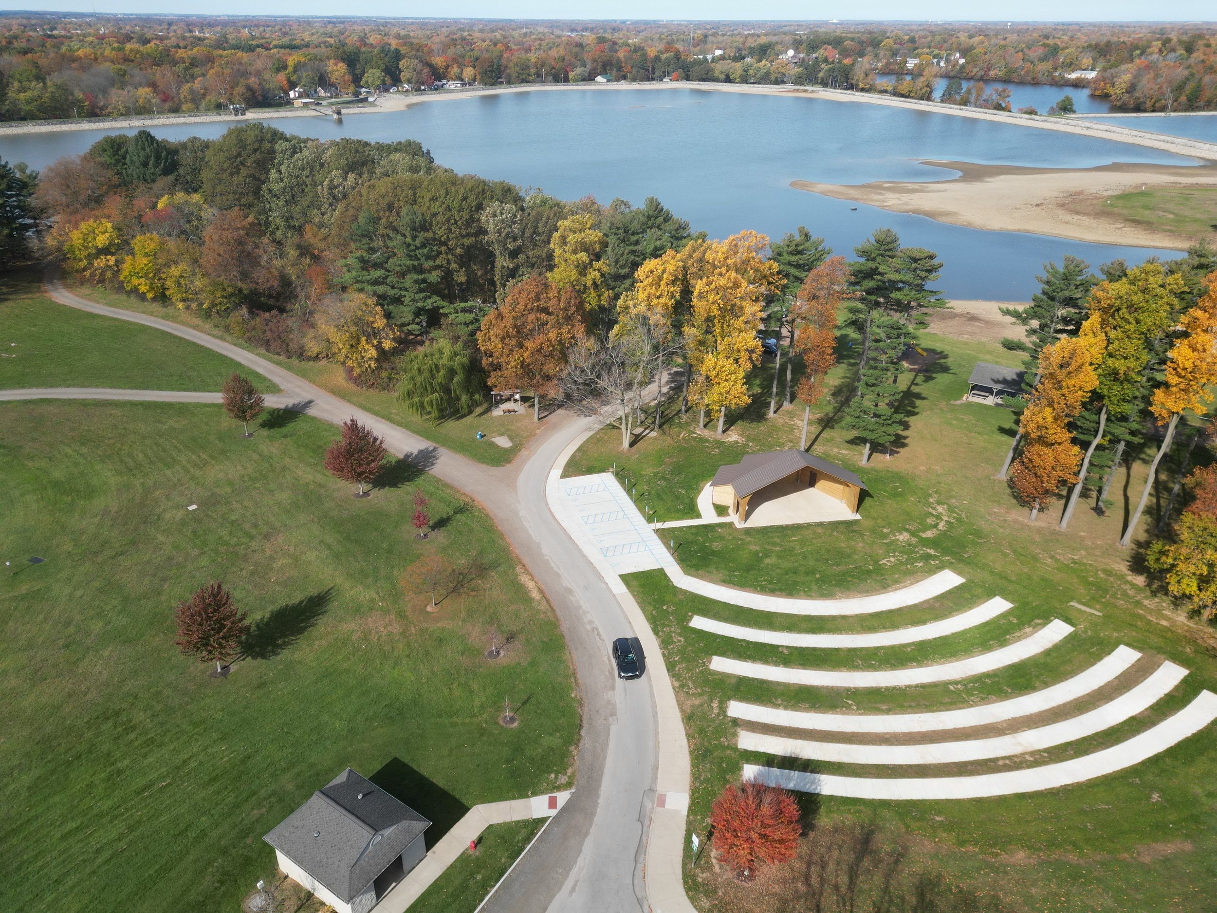 An aerial view of a park with a large body of water, surrounded by vibrant autumn trees, a small pavilion, and winding walkways.