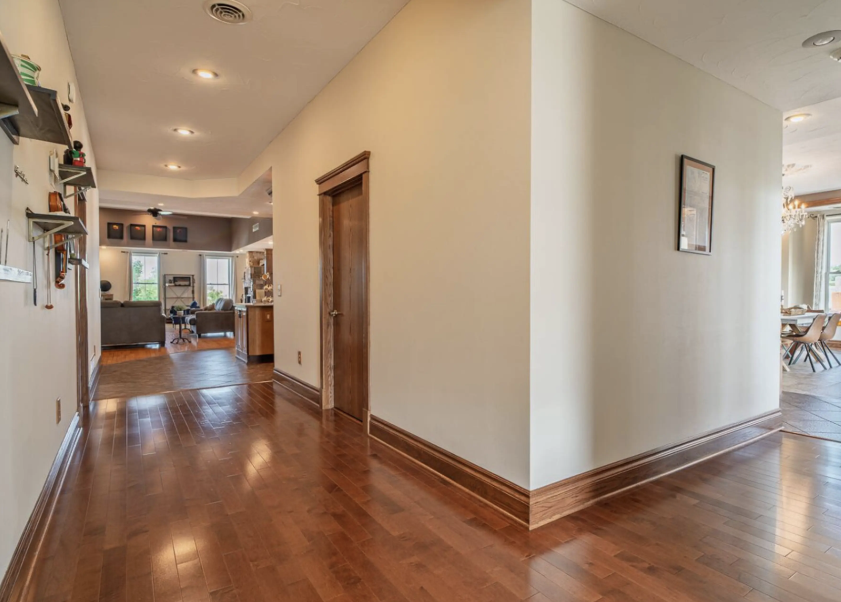 A hallway with hardwood floors and white walls leading to a living room with windows and furniture, with a partial view of a dining area and decorative shelving on the left wall.
