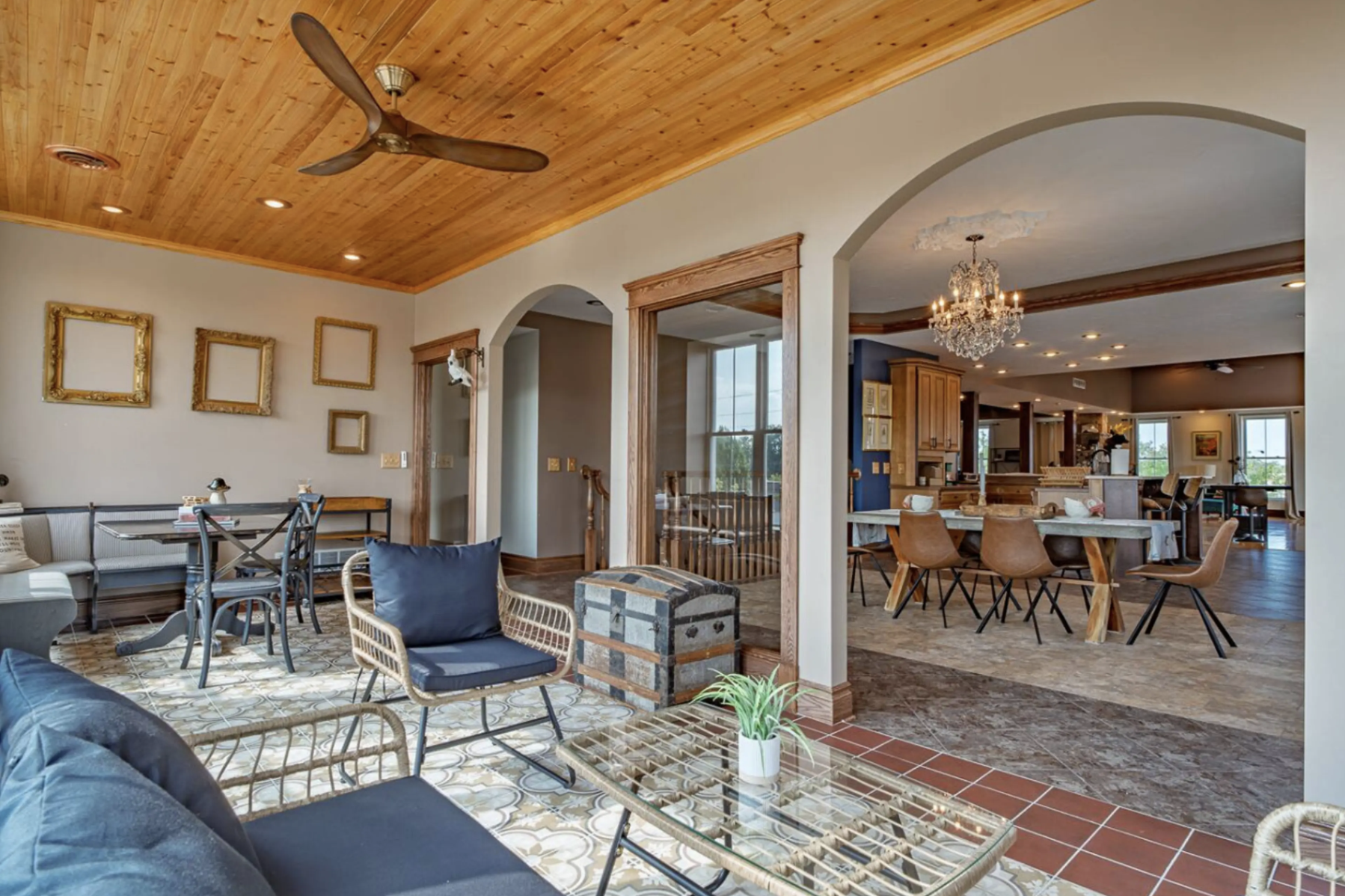 A spacious open-concept living and dining area with a wooden ceiling, ceiling fan, chandelier, and large windows. The space is decorated with modern and vintage furniture, including a round dining table with chairs, a sofa, armchair, and framed artwo