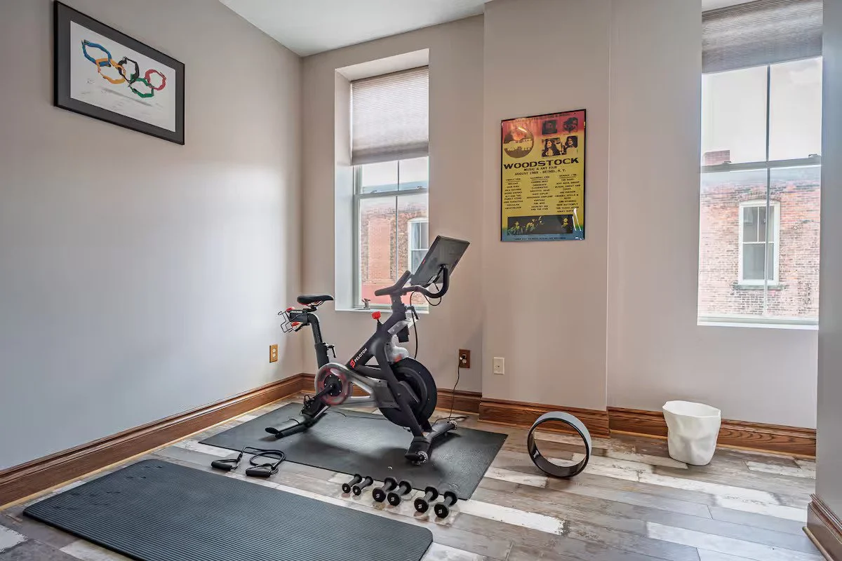 Home exercise room with Peloton bike, yoga mats, small weights, and screen, near windows with view of brick buildings.