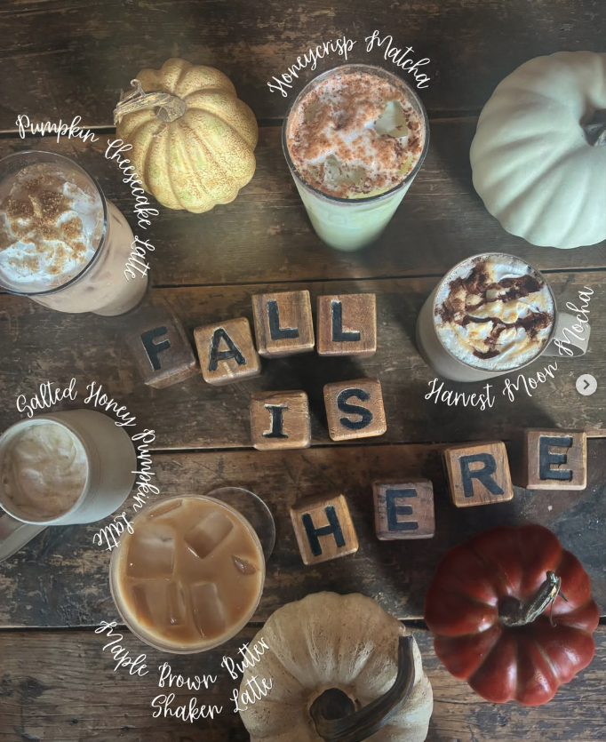 A rustic wooden table with seven different pumpkin-themed drinks labeled with white handwritten text. The drinks include Pumpkin Cheesecake Shake, Honeycrisp Matcha, Harvest Moon Toffee, Salted Honey Butterscotch, Apple Brown Butter Shaken Latte, and two other drinks. Decorations include a gold pumpkin, a white pumpkin, and a red tomato. In the center, wooden blocks spell out "FALL IS HERE."