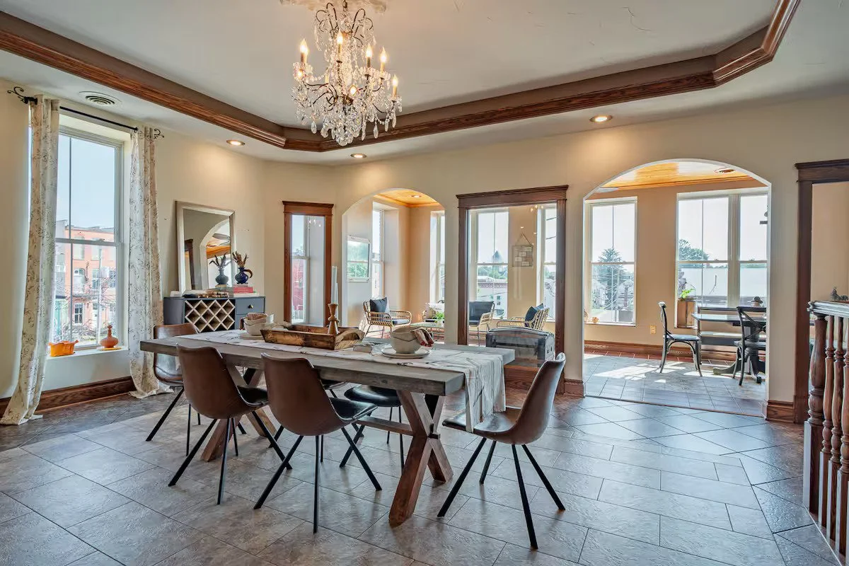 Bright dining area with a rustic wooden table, six modern brown chairs, a chandelier hanging from a tray ceiling, large windows with floral curtains, and a sunlit sitting nook with chairs and a small table.
