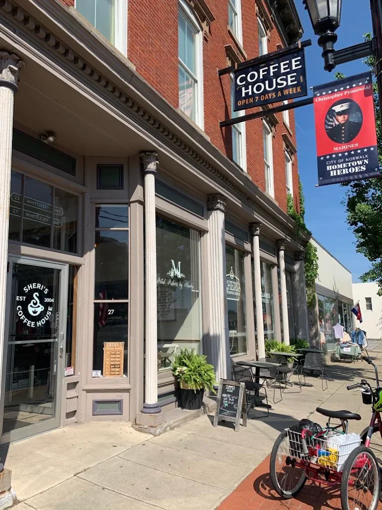 The exterior of Sheri's Coffee House, a coffee shop with large windows, white columns, and outdoor seating. A sign indicates it is open seven days a week. There are potted plants and a chalkboard sign on the sidewalk, along with a bike parked nearby.