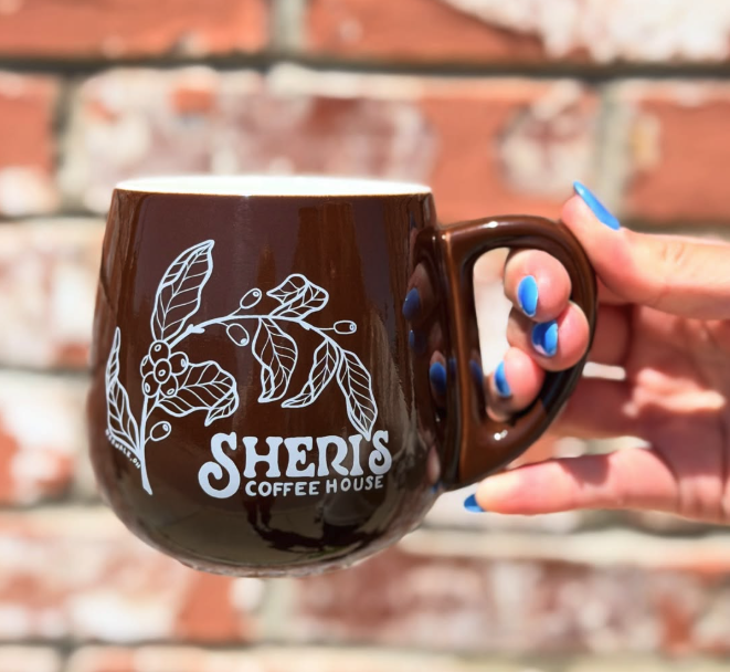 A person with blue painted nails holds a large brown coffee mug with a white floral design and the text 'Sheri's Coffee House' written on it, against a blurred brick wall background.