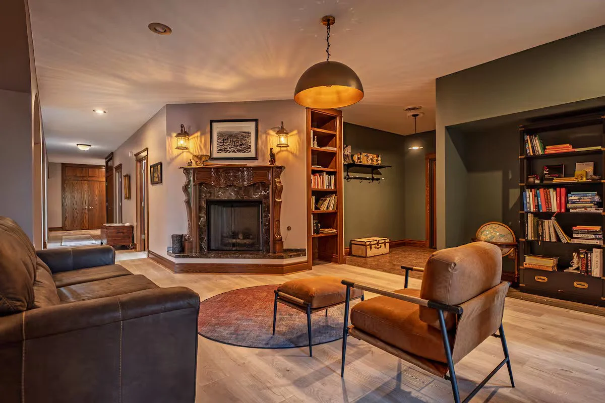 Living room with a fireplace, bookshelf, leather armchair, and sofa, decorated with framed photos and warm lighting.