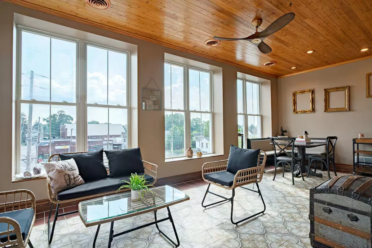 Bright living room with large windows, wooden ceiling, ceiling fan, black cushioned furniture, and decorative picture frames on the wall.