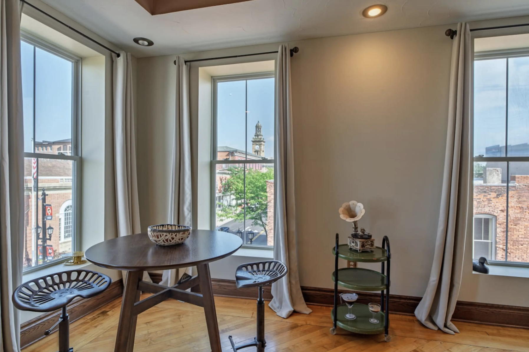 Sunlit dining area with three large windows showing a cityscape with a clock tower, brick buildings, and green trees. A round dining table with black chairs and a decorative bowl sits near the windows. Cream curtains frame the windows, and an antique