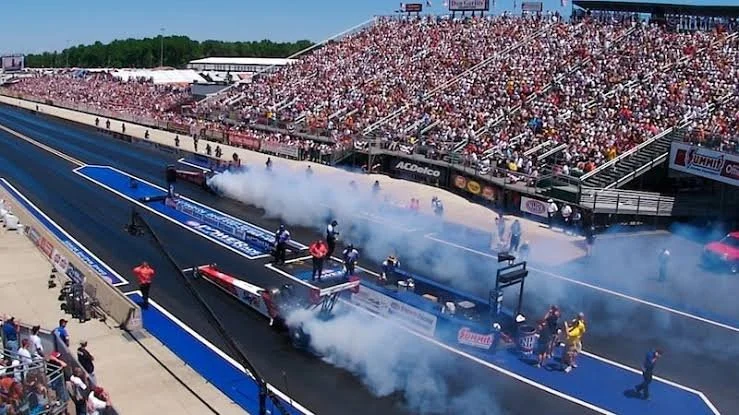 Drag racing track with cars and crew members preparing, with smoke from engines, a packed grandstand of spectators, and a clear blue sky.
