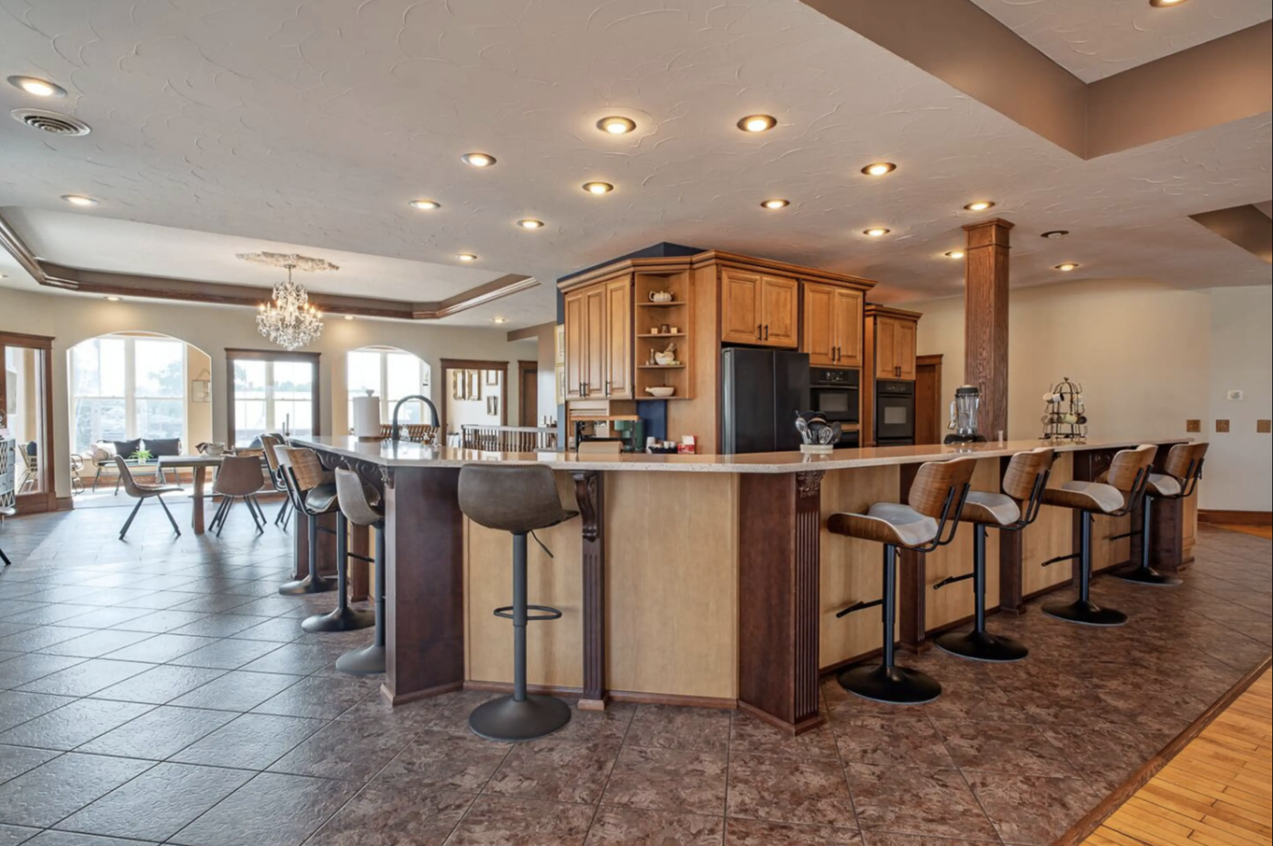Spacious kitchen and dining area with a large curved island, bar stools, wooden cabinets, and a chandelier, with large windows and tiled flooring.