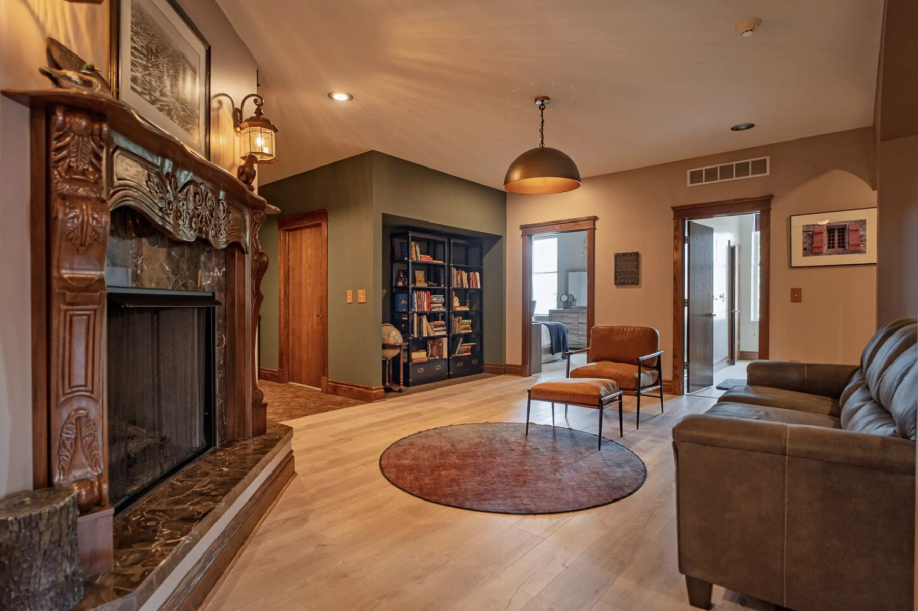 A cozy living room with a wooden fireplace, a black bookshelf filled with books, and a brown armchair with an ottoman. The room has hardwood floors, warm-colored walls, and a circular rug. There are two doorways leading to other rooms, with one havin
