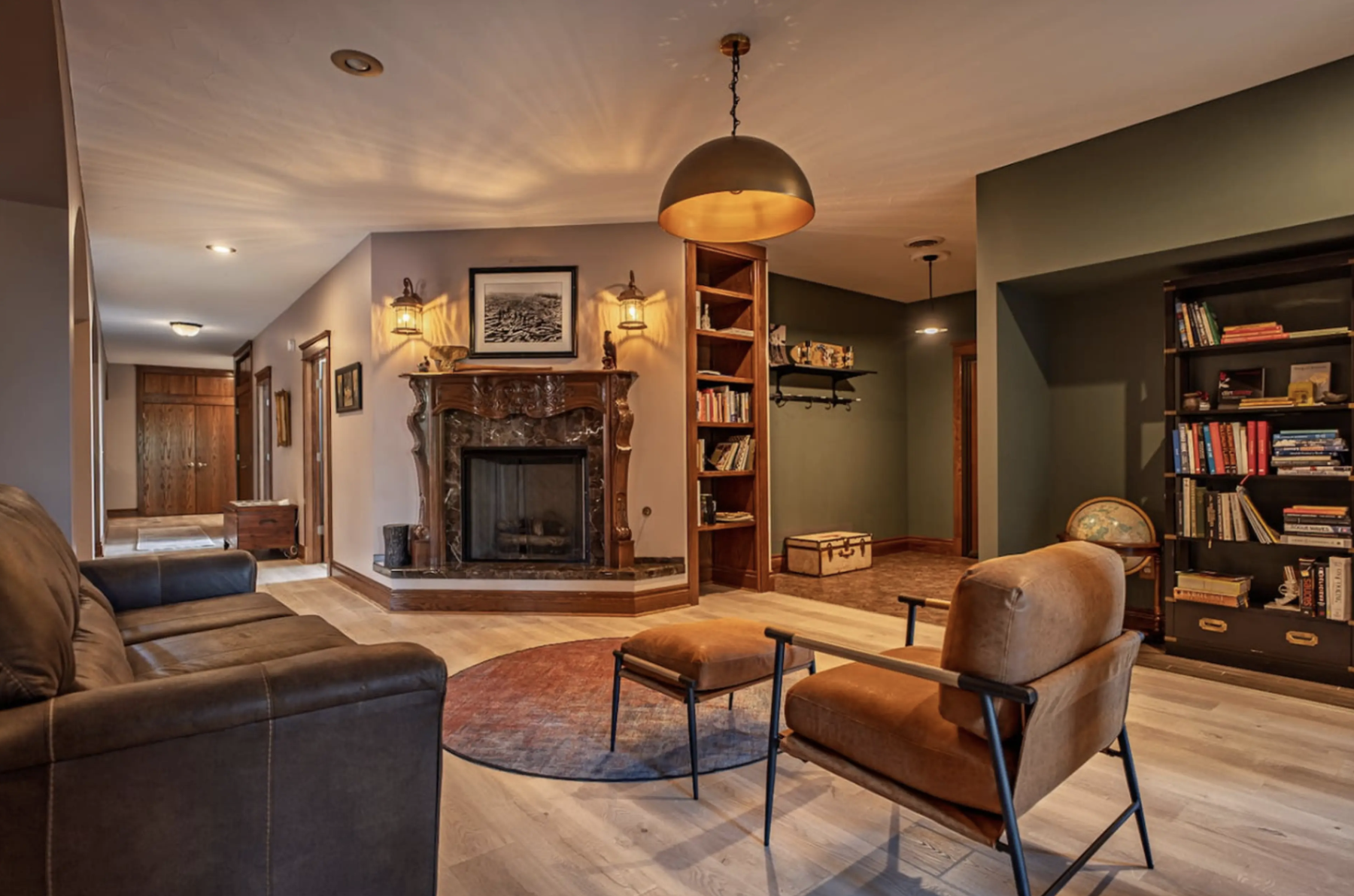 Living room with a leather sofa, an armchair, and a round rug. A fireplace with a decorated mantel is on the wall, with framed artwork and wall-mounted lamps. There are bookshelves and a globe, with warm lighting creating a cozy atmosphere.