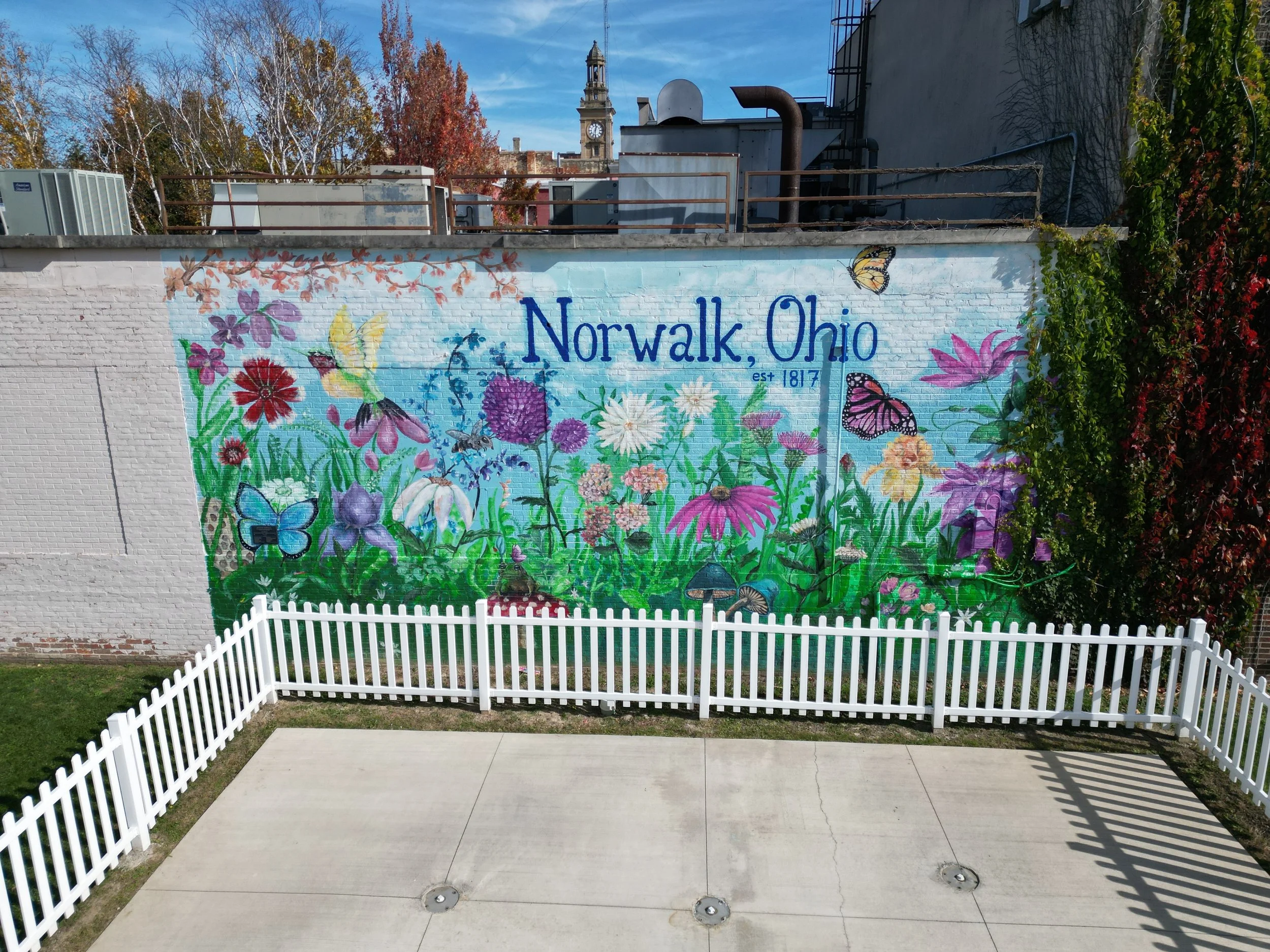 Colorful mural on a brick wall in Norwalk, Ohio, featuring flowers, butterflies, and the text "Norwalk, Ohio est 1817" with a white picket fence in the foreground.