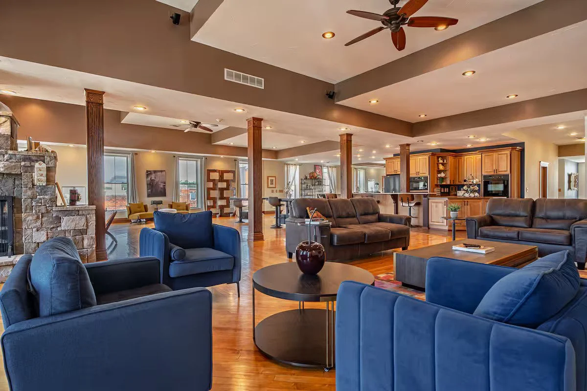 Spacious open-concept living room with hardwood floors, multiple sofas and chairs, a stone fireplace, large windows, and a wooden kitchen with bar seating in the background.
