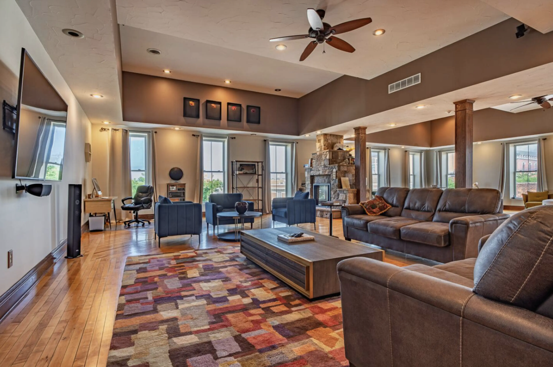 Spacious living room with large windows, hardwood floors, leather sofas, armchairs, a stone fireplace, ceiling fans, and a colorful geometric rug.