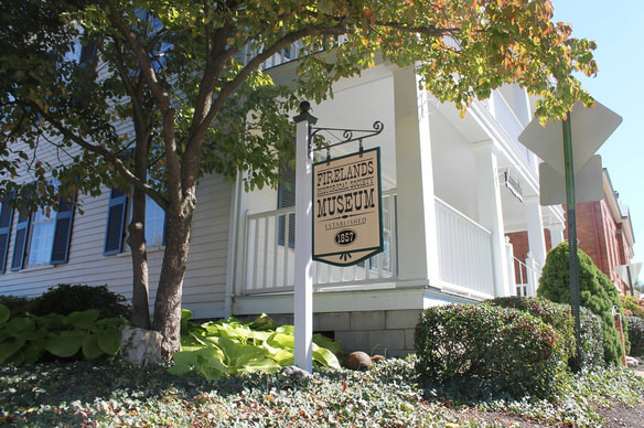 Sign for Firelands Museum outside a white building with a porch, surrounded by trees and bushes.
