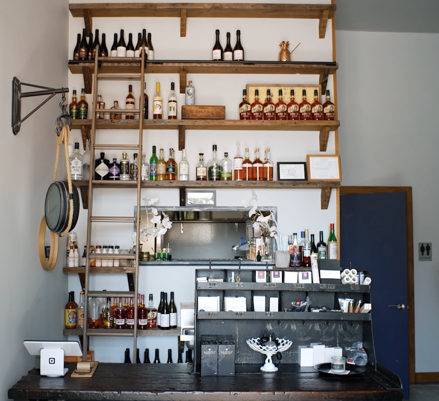 Bar area with wooden shelves holding various bottles of alcohol, a ladder, and decorative items. A small counter with additional bottles, equipment, and a blackboard to the side.
