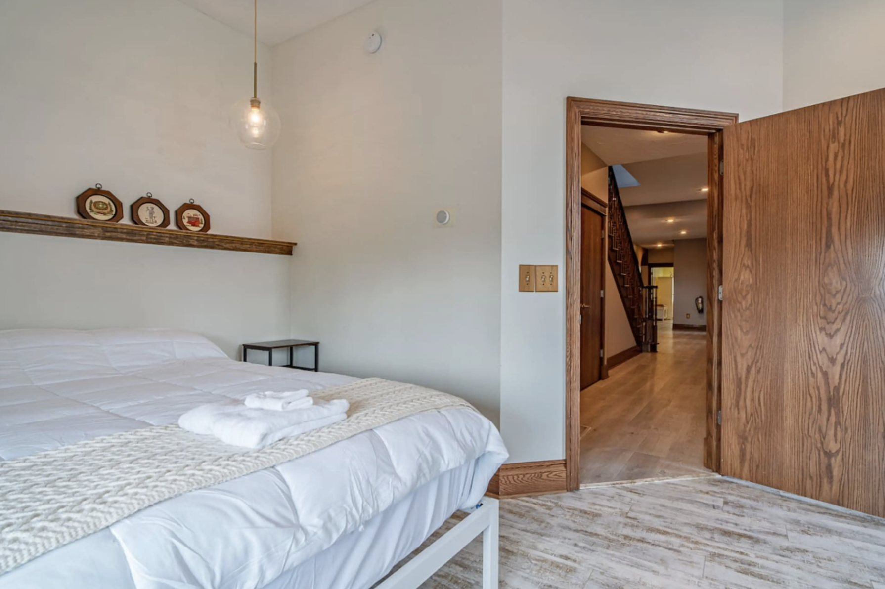 A cozy bedroom with a neatly made bed, white linens, and folded towels on top, a black nightstand, and decorative plates on a wooden shelf above the bed. The room has white walls, a hanging light bulb, and a doorway leading to a hallway with wooden f