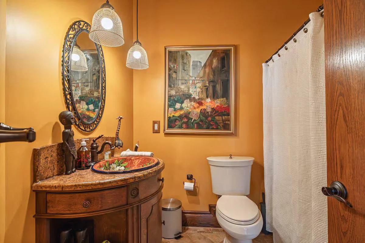 A bathroom with yellow walls, a wooden door, a toilet, a wooden vanity with a decorative sink, a mirror, hanging pendant lights, a painting on the wall, a white curtain, and various decorative items on the vanity.