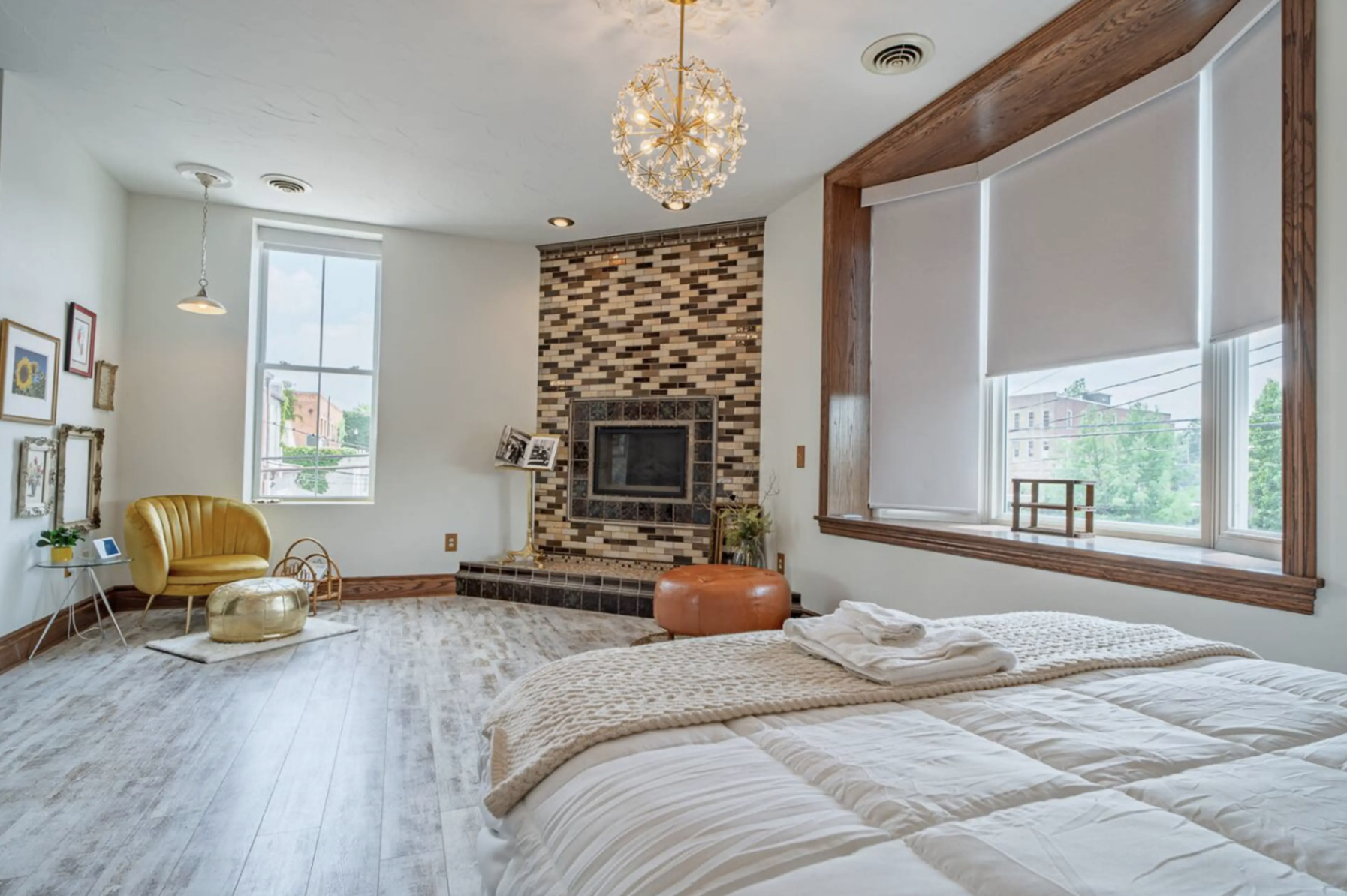 A cozy bedroom with large white beds, folded towels, and a wooden window ledge with blinds. The room features a fireplace with a brick and tile surround, a modern chandelier, and artwork on one wall. There's a yellow chair with a matching ottoman, a 